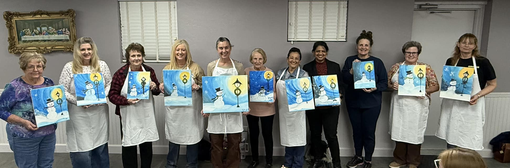 Paint night attendees pose holding their paintings of snowmen in lamplight